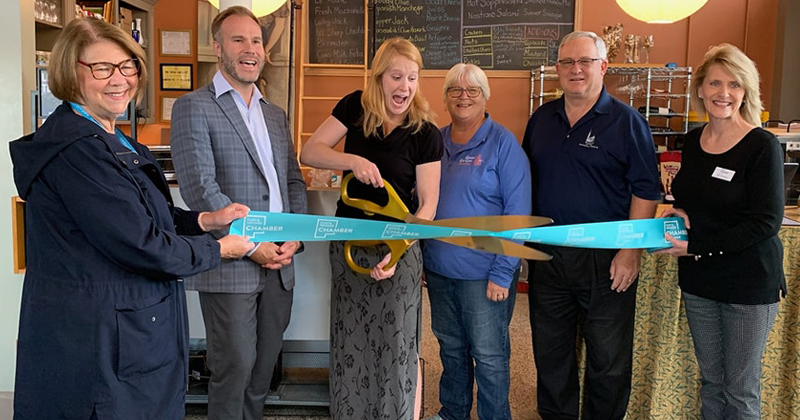 Owner of Queens' Provisions cuts a ribbon with large scissors to open her new business. Two employees are at her side along with three Chamber members.