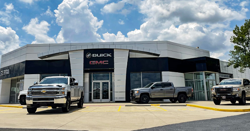 View of outside building of Serra Buick GMC car dealership with three silver trucks on display