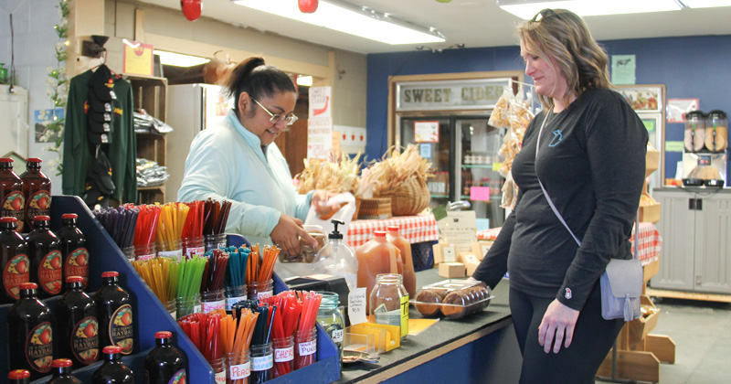 Inside Almar Orchards fruit market, a cashier checks out a customer who is purchasing cider and donuts surrounded by a display of bottles of hard cider and flavored honey sticks for sale.