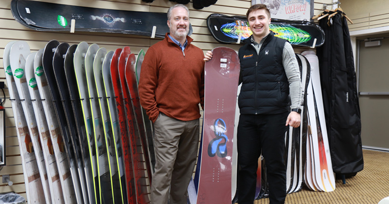 Tony Shumaker stands next to his son, Garret who is holding one of snowboards they sell in their ski shop. Behind them is a wall lined with snowboards.