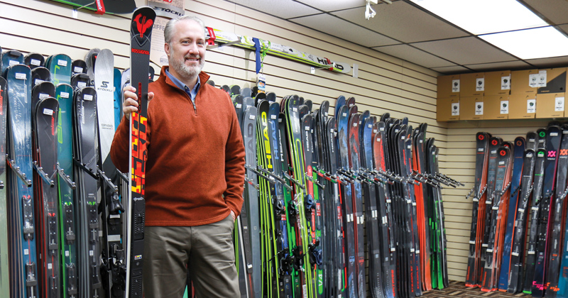 Ski Shop owner, Tony Shumaker, stands in front of a wall lined with snow skies. Shumaker is holding a black set of skies with yellow and red graphics.