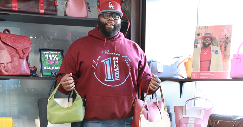 Miracle McGlown, wearing a 1:Eleven logo red hoodie holds four of his leather bags on his arms in front of his shelves displaying more of his leather bags inside Shops on Saginaw.