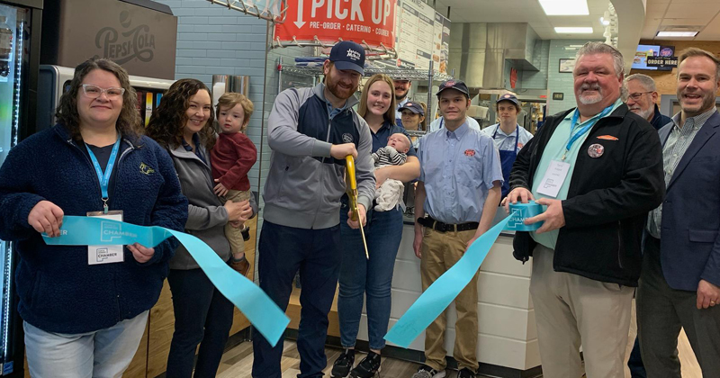 The owner of Jersey Mike's is joined with his wife and employees inside his newest Jersey Mike's sandwich shop to cut a  grand opening ribbon.