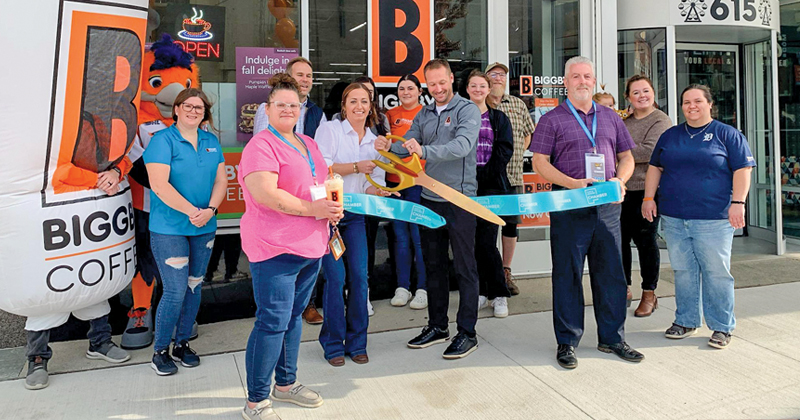 A group of people are lined up in front of the Biggy Coffee Shop holding a ribbon while a man and woman in the middle cut the ribbon with large scissors.