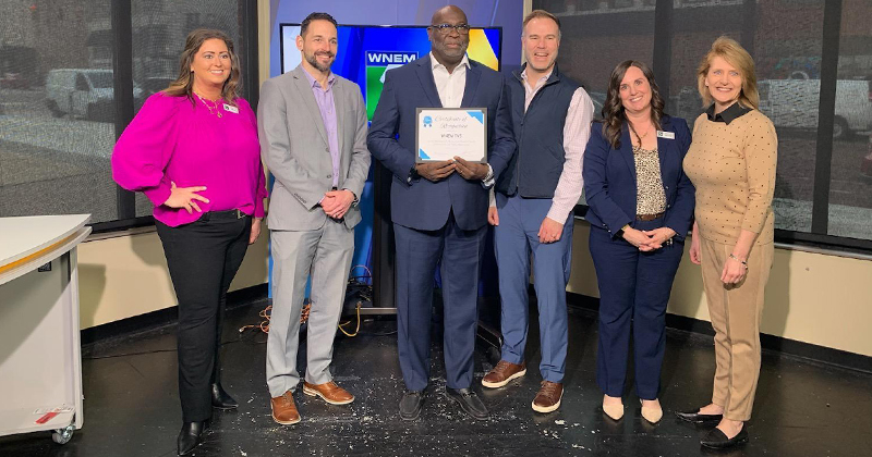 A chamber employee joins four employees of WNEM-TV inside their studio to celebrate their 70th anniversary. One of the employees holds a certificate to commemorate the milestone.