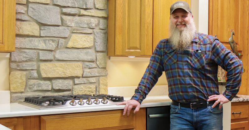 A man with a long beard dressed in jeans and plaid shirt and baseball cap leans against a quartz countertop of a mock kitchen.