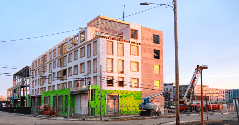 Exterior view of the L-shaped construction of the LiveWell on Harrison YMCA building. The exterior walls, minus windows, are in place in the southern end of the structure. The norther end of the structure is framed in steel beams. Construction crews operate forklifts to work on the exterior.