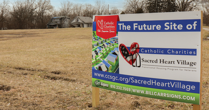 An empty city lot with a large sign stating it would be the future site of Sacred Heart Village.