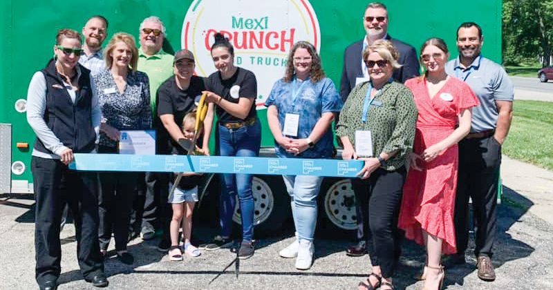 Staff and chamber members line up in back of a large blue ribbon for a ribbon cutting in front of the green Mexi Crunch food truck.