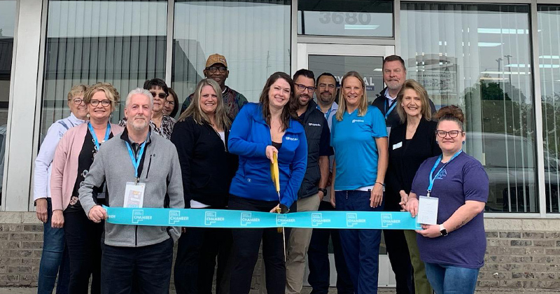 Staff and chamber members line up in back of a large blue ribbon outside the offices of Fyzical Therapy for a ribbon cutting