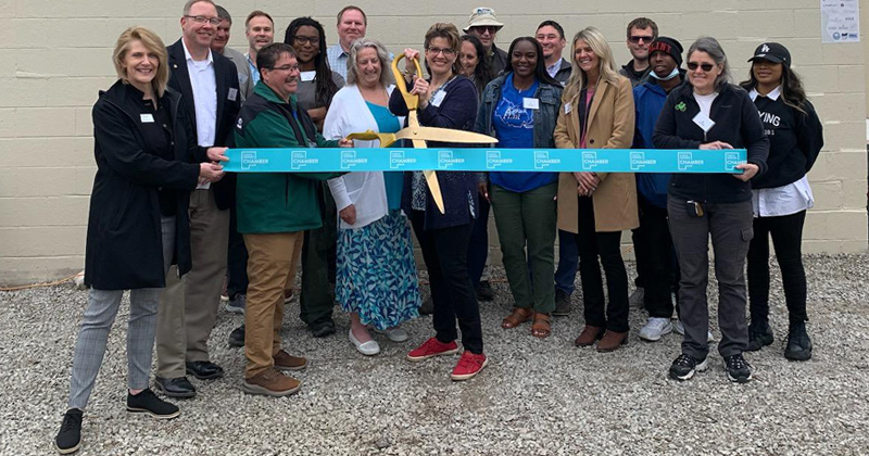 Friends of the Fllint River volunteers, staff and chamber members line up in back of a large blue ribbon for a ribbon cutting 