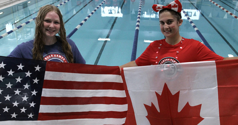 Two young women swimmers standing in front of the pool hold up the flag they are representing. The United States on the left and Canada on the right.