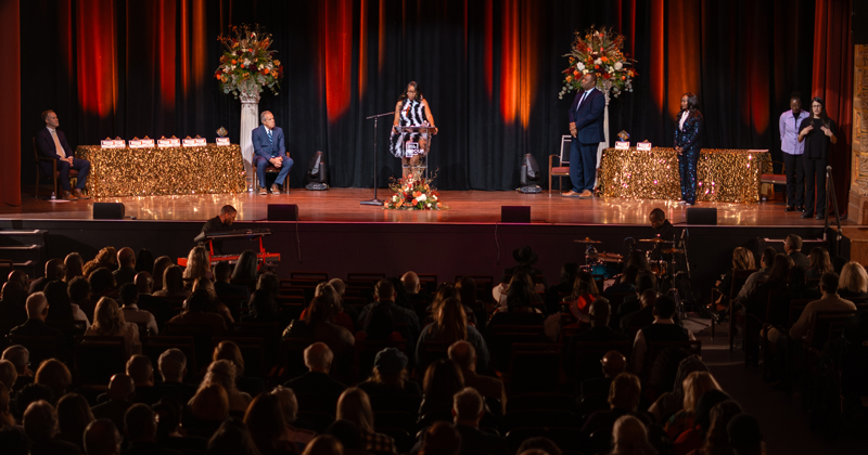 View of the stage at the Capitol Theatre from the back. On the stage a woman makes a statement at the podium in the middle of the stage. On each side is a table with awards. The theatre is filled with attendees.