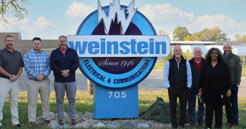 Owners and management of Weinstein Electric (three men on the left and three men and one woman on the right) line up in front of the company sign outside their building.