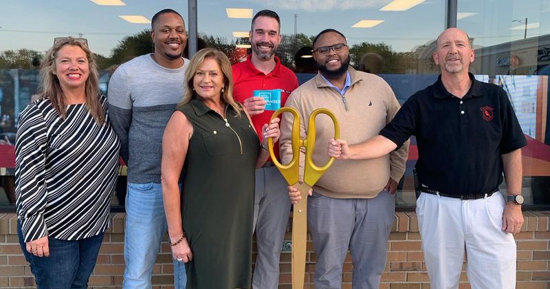 Six employees line up outside the company building for a ribbon cutting. Three employees hold gold oversized scissors that will be used to cut the ribbon.