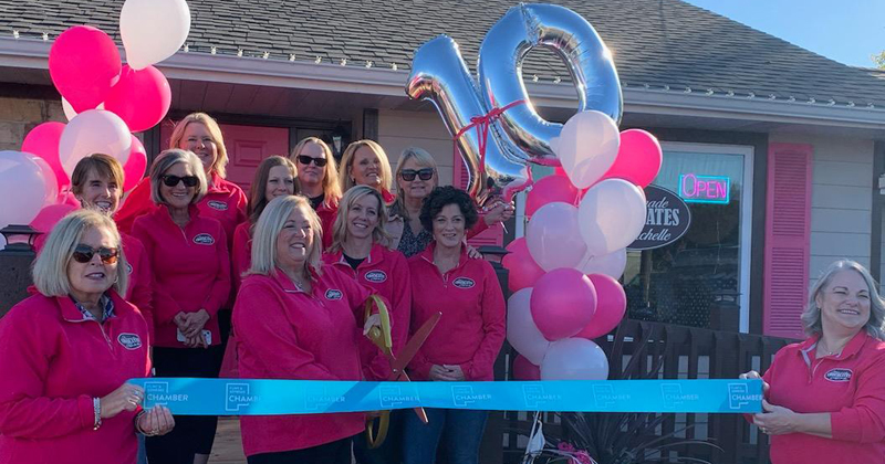 Ten women dressed in pink jackets line up in three rows outside the store for a ribbon cutting. Pink and white balloons flank both sides of the women. Two additional women also dressed in pink jackets hold a blue ribbon with the chamber logo repeated across it. The owner stands in the middle and holds gold oversized scissors open to cut the ribbon.