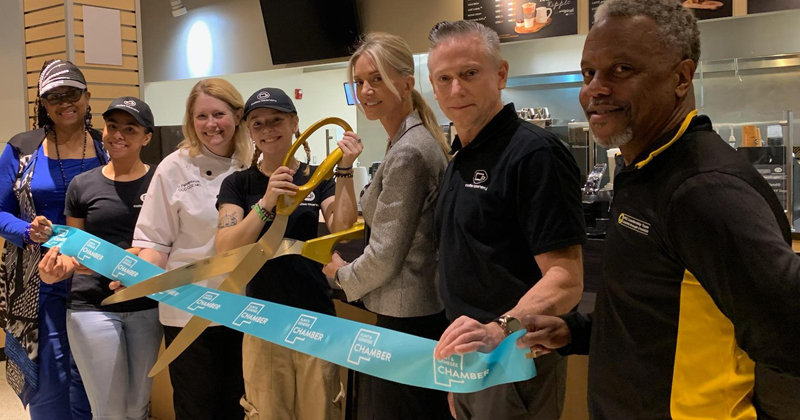 Seven people line up in front of the pastry display counter for a ribbon cutting. The pastry chef in her white coat, third from the left, helps hold up the ribbon for a culinary student who is to her right. The student holds gold oversized scissors getting ready to cut a large blue ribbon with the chamber logo repeated across it.