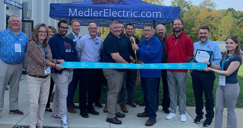 Employees line up outside the company building for a ribbon cutting alongside two owner who both hold gold oversized scissors getting ready to cut a large blue ribbon with the chamber logo repeated across it. A blue canopy with Medler Electric.com is in the background.