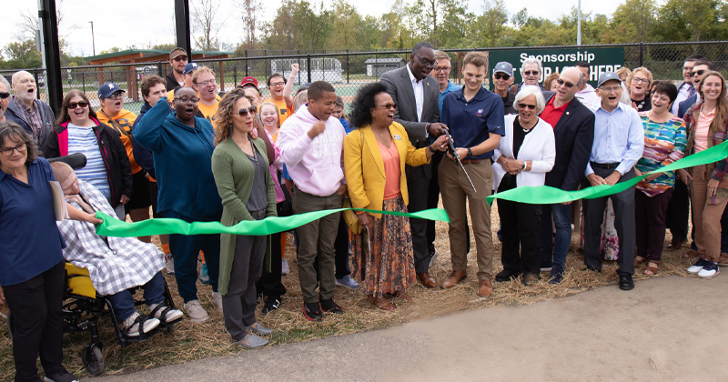 A crowd of people line up outside the a new handicap accessible ballfield for a ribbon cutting. The Michigan Lt. Governor and the young man who began the project hold oversized scissors getting ready to cut a large green ribbon.