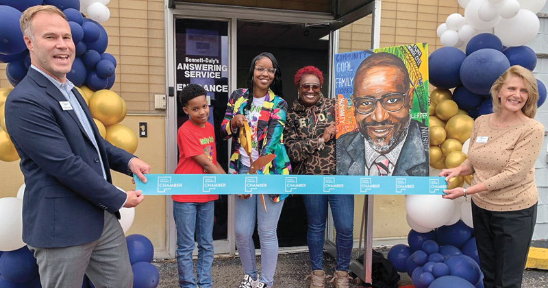 Employees and Chamber staff line up outside the Healthy Just Feels Right office to cut a long ribbon with the chamber logo printed on it. The owner is in the middle and has large scissors to cut the ribbon. To the right of the owner is a poster with a headshot of the late BB Nolden.