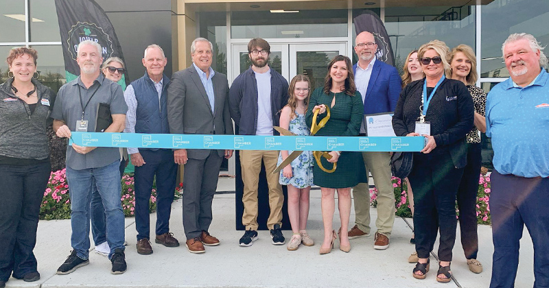 Employees line up outside the company building for a ribbon cutting alongside the owner who holds gold oversized scissors getting ready to cut a large blue ribbon with the chamber logo repeated across it.