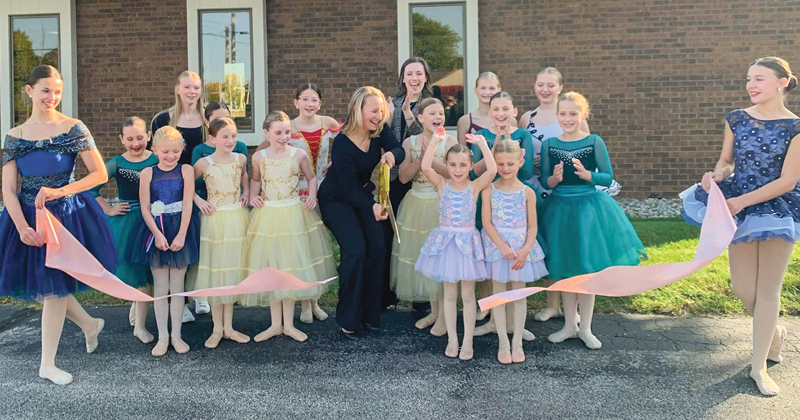 Young dance students dressed in the tutus line up outside the dance studio building for a ribbon cutting alongside the owner who holds gold oversized scissors getting ready to cut a large pink ribbon.