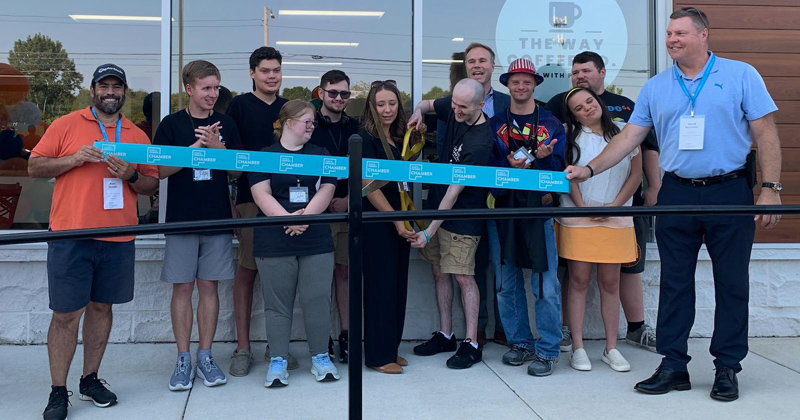 Young employees of The Way Coffee line up outside the company building for a ribbon cutting alongside the owner who holds gold oversized scissors getting ready to cut a large blue ribbon with the chamber logo repeated across it.