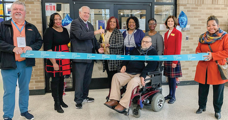 Employees line up outside the bus station for a ribbon cutting alongside the owner who holds gold oversized scissors getting ready to cut a large blue ribbon with the chamber logo repeated across it.