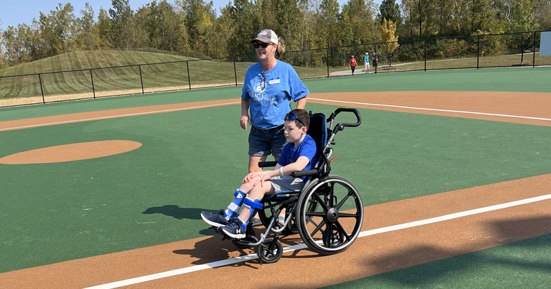 A woman pushes a young boy in his wheelchair around an accessible baseball field.