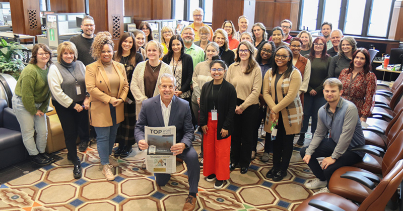 A man holding a newspaper clipping announcing the organization's award kneels in front of four rows of staff members.