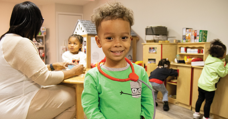 A little boy in a green shirt with a gray monster on the label wears a pretend stethoscope around his neck. In the background, his child care worker sits at a small table and plays tea with another young child.