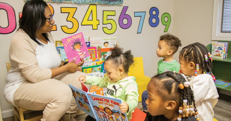 A child care worker sits on a child's chair in front of four young children who gather around to listen to the book she is holding.