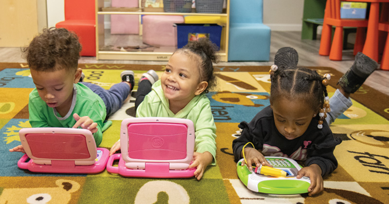 Three young children are spread out on a large rug playing with pretend computers.