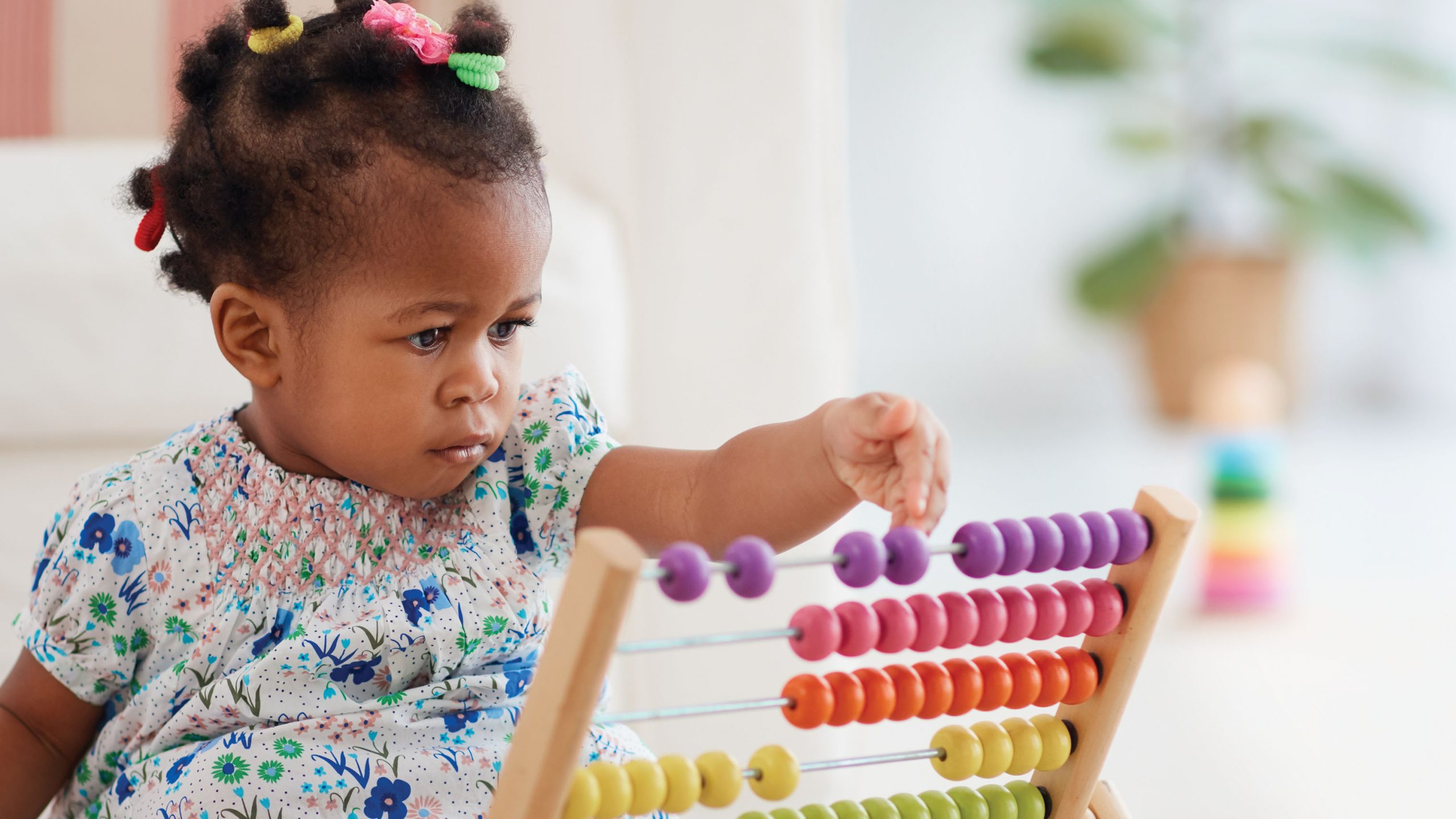 A little girl, no older than two, dressed in a colorful dress, pushes the a purple bead on a colorful abacus.