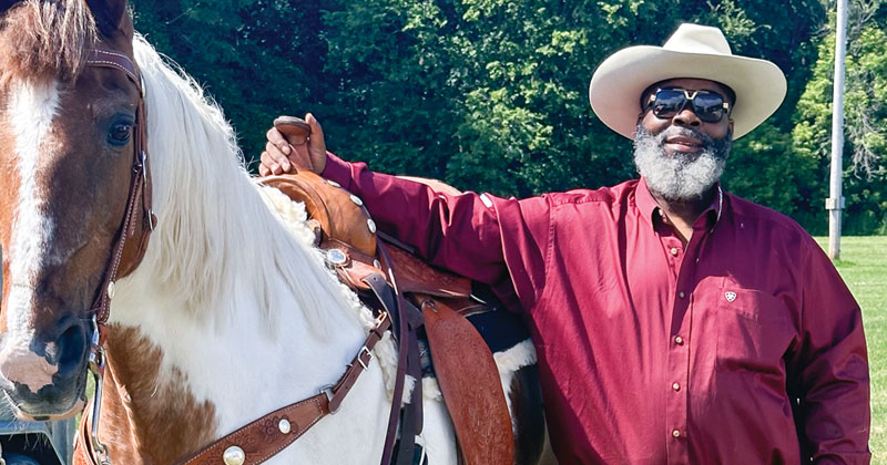 An older man with a gray beard wearing large sunglasses and a cowboy hat stands next to his white and brown horse.