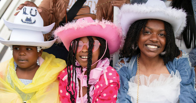 Two young girls sit next to their mother on the bleachers at a rodeo. The girl on the left is wearing a yellow rain poncho and two white cowboy hats. The young girl next to her is wearing a pink shirt underneath a pink polka-dot raincoat. a pink cowboy hat and pink heart-shaped glasses. Their mother is wearing a blue denim jacket and white lace cowboy hat.