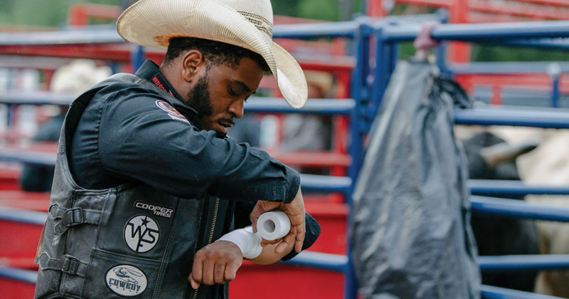 A Black cowboy wearing a black leather vest and cream cowboy hat, wraps his wrist with tape in preparation of riding a bronco.