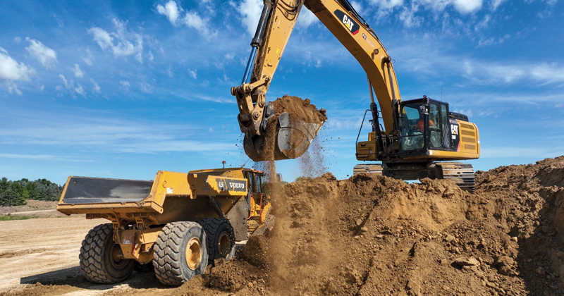 An large excavator transfers a bucket of soil into the back of  a waiting dump truck.