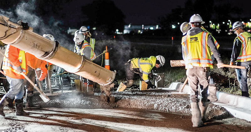 Crews work at night under lights to pour cement for a roadway.