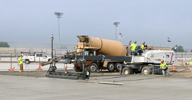 A cement mixer dumps fresh cement from the mixer as workers use long handled rakes to spread the concrete
