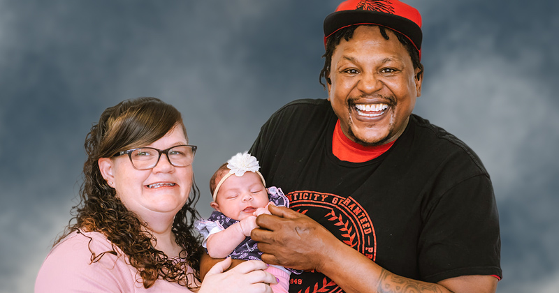 A mother in a pink dress and father wearing a black collegiate t-shirt and red baseball cap hold their baby daughter wearing a flower dress and white headband with flower attached.