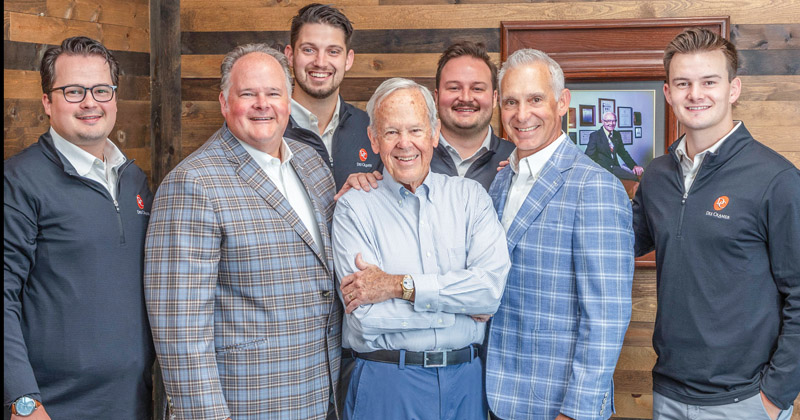 A generation photo showing seven current employees of Dee Cramer standing in front of a photo of the deceased founder.