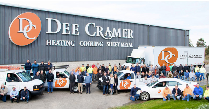 A landscape staff photo of employees at Dee Cramer. The staff re mingled among three company trucks, a company semi and company car all in the parking lot in front of the warehouse with their company name  and logo visible on the building.
