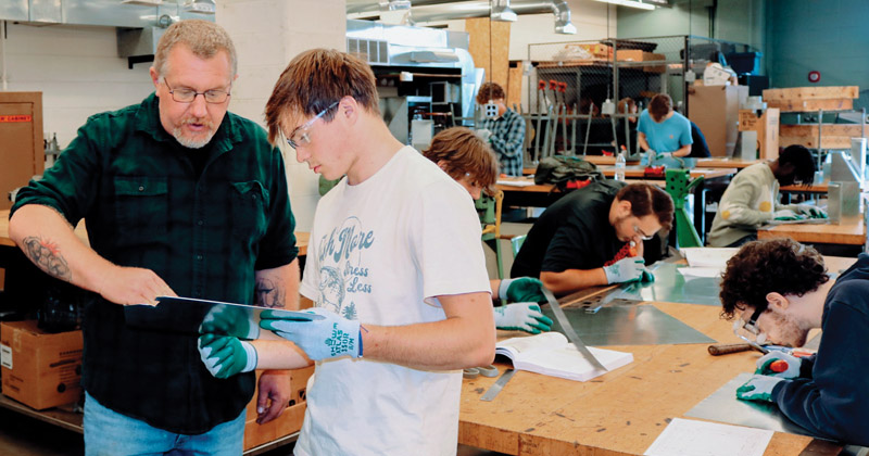 An instructor assists a student with his metal work in an HVAC class while other students sit at a table and work on their projects.