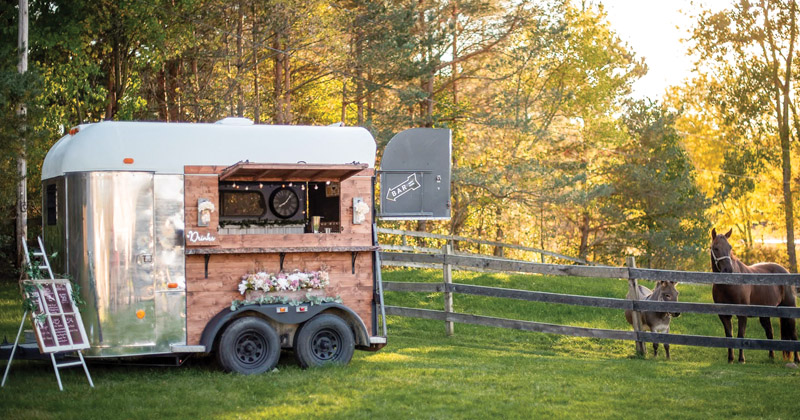 A converted horse trailer to be a bar on wheels is parked in a horse corral. A sign showing drinks available is to the left of the trailer. The trailer side windows are open with a shelf to order drinks. Horses look onl.