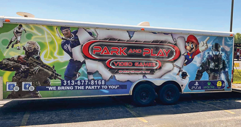 A trailer, almost the size of a semi-trailer, is parked in a parking lot. The trailer is wrapped with colorful images of various vide game characters such as Mario Brothers.
