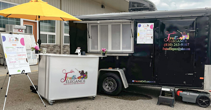 A black food truck is parked in a parking lot. Two double sets of windows on the side give easy access into the trailer. A separate rolling cart shaded by a bright yellow umbrella is ready to serve people. An easel with beverage menu is set up next to the rolling cart.
