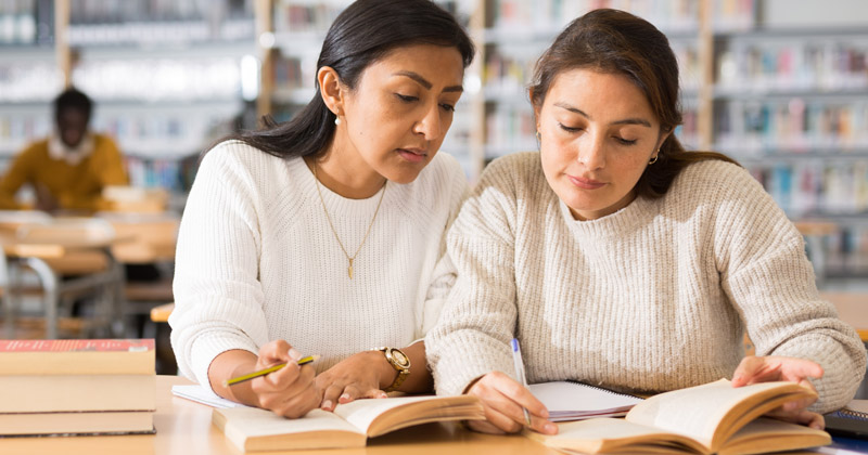 Two women, both wearing long, white sweaters, are sitting at a table inside a library. Books are open in front of them both. The woman on the left is overlooking and instructing the other woman who is pointing to something in the book.