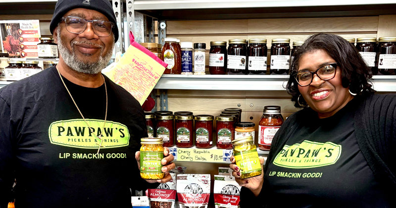 Husband and wife stand on ends of tall display of their pickles they make and sell.